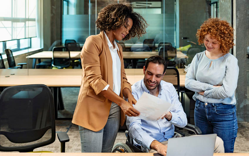 One Businessman on a wheelchair sitting at the table with his two female colleagues standing next to him in the office 